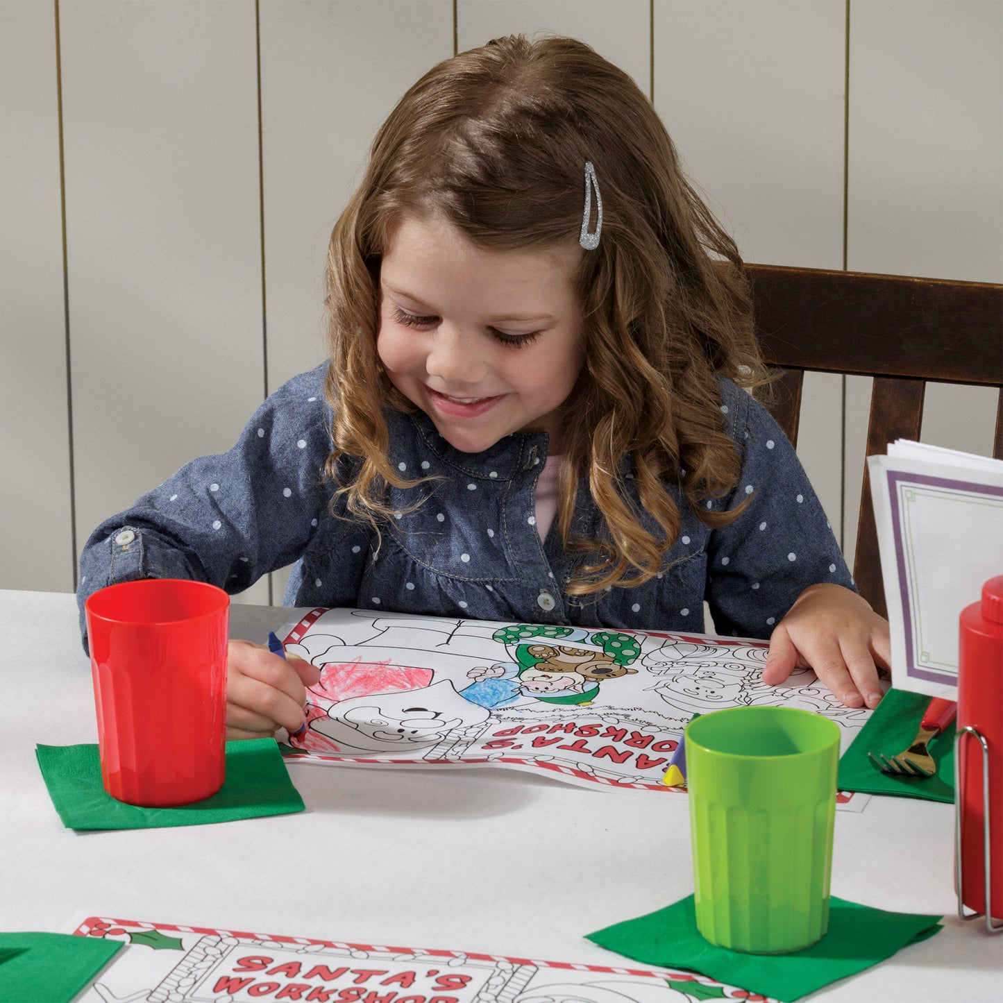 Young girl coloring with crayons at a table with Christmas-themed coloring pages and cups.