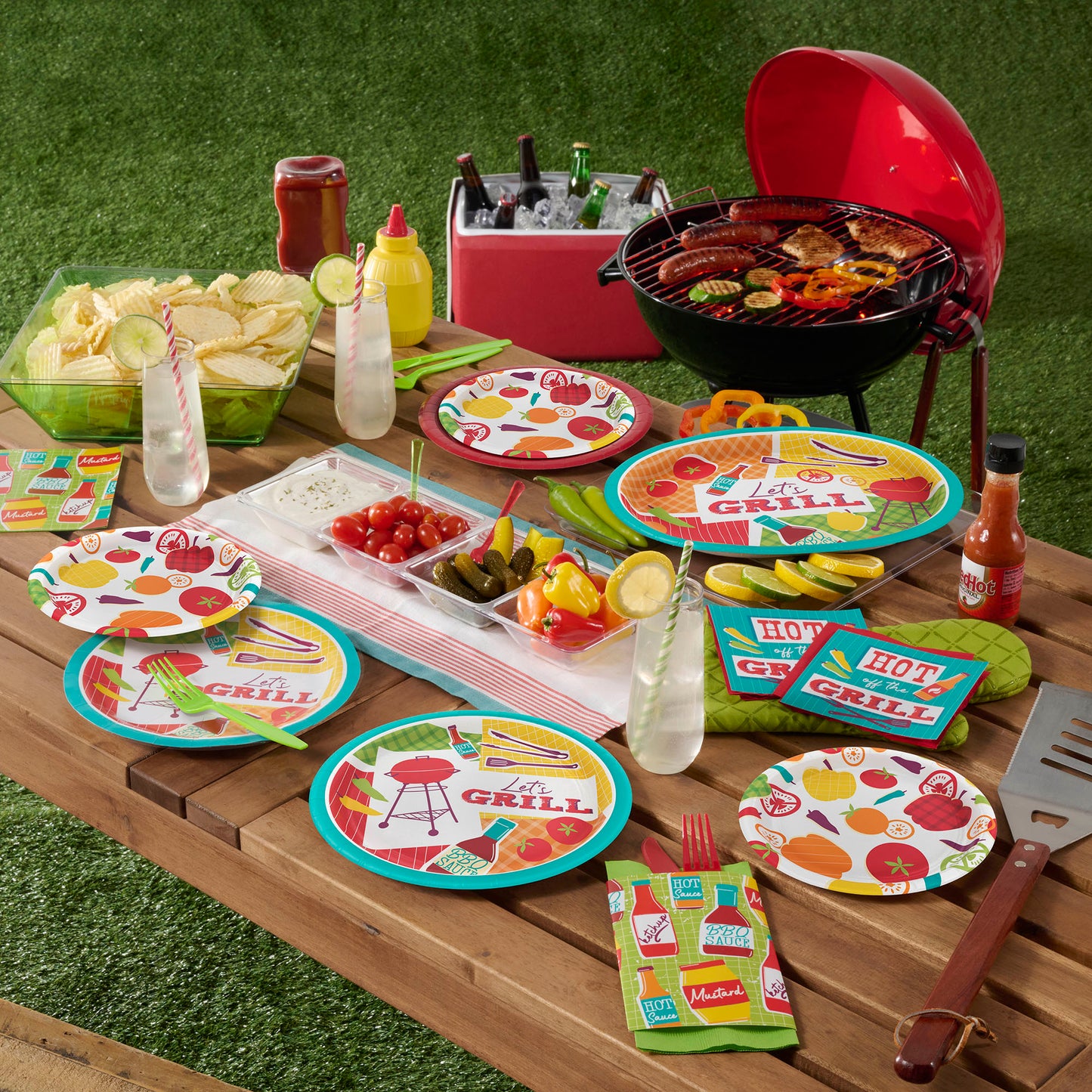 Outdoor barbecue setup with plates and napkins on a wooden table with food and a grill.