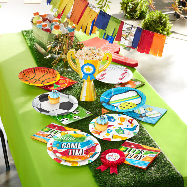 Sport themed party supplies displayed on a green table with basketball, baseball, soccer, and pickleball-themed plates and napkins