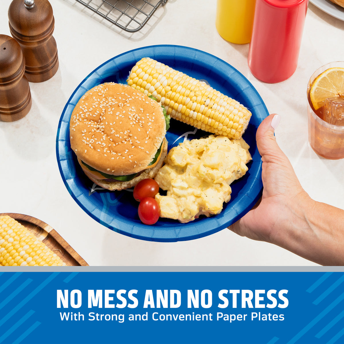 A hand holds a blue paper plate with a hamburger, corn on the cob, mashed potatoes, and cherry tomatoes on a kitchen counter. Condiment bottles, pepper mills, and a drink are visible in the background, with a blue banner at the bottom reading 'NO MESS AND NO STRESS With Strong and Convenient Paper Plates'.