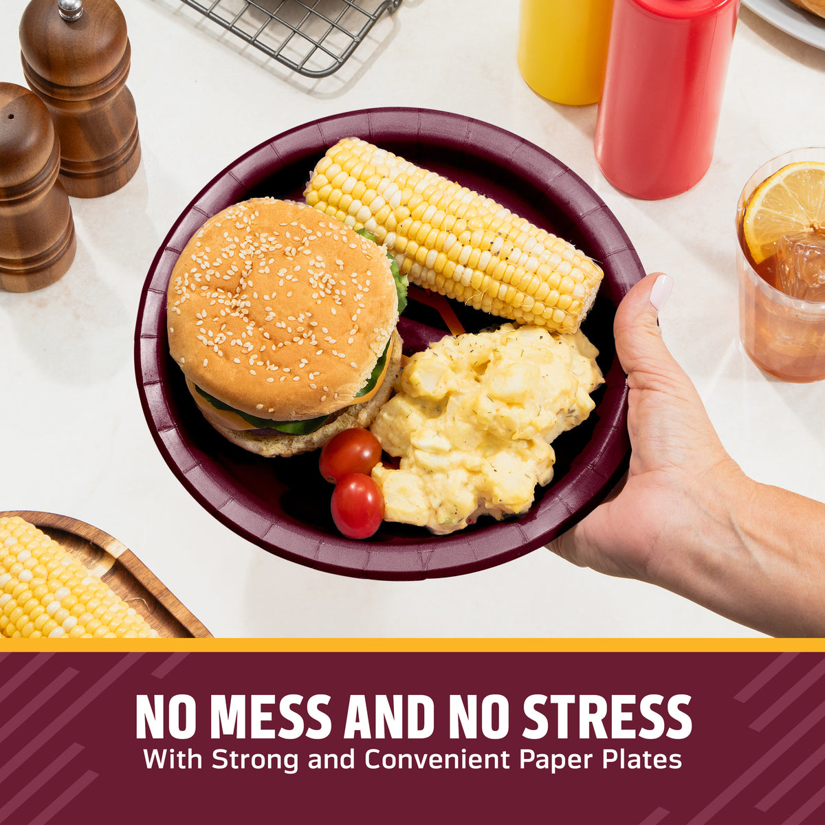 A hand holds a dark burgundy red  Washington Commanders paper plate containing a hamburger, corn on the cob, potatoes, and cherry tomatoes on a kitchen counter. Condiment bottles, pepper mills, and a drink are visible in the background, with a text banner at the bottom reading 'NO MESS AND NO STRESS With Strong and Convenient Paper Plates'.