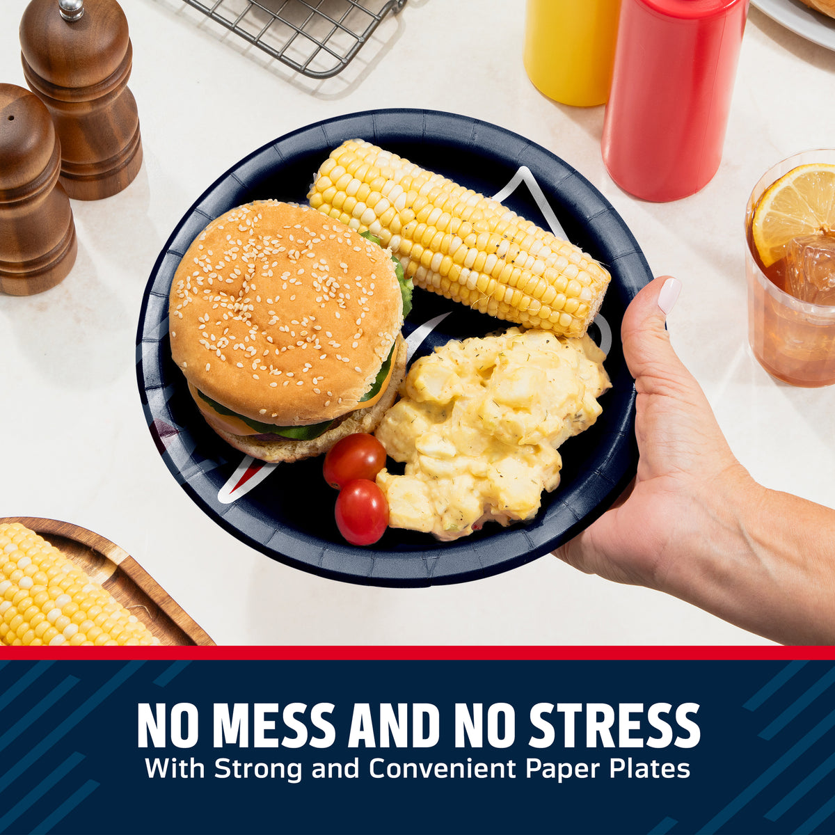 A hand holds a black paper plate with a hamburger, corn on the cob, potatoes, and cherry tomatoes on a white countertop. Condiment bottles, pepper mills, and a drink are visible in the background, with a dark blue banner at the bottom reading 'NO MESS AND NO STRESS With Strong and Convenient Paper Plates'.