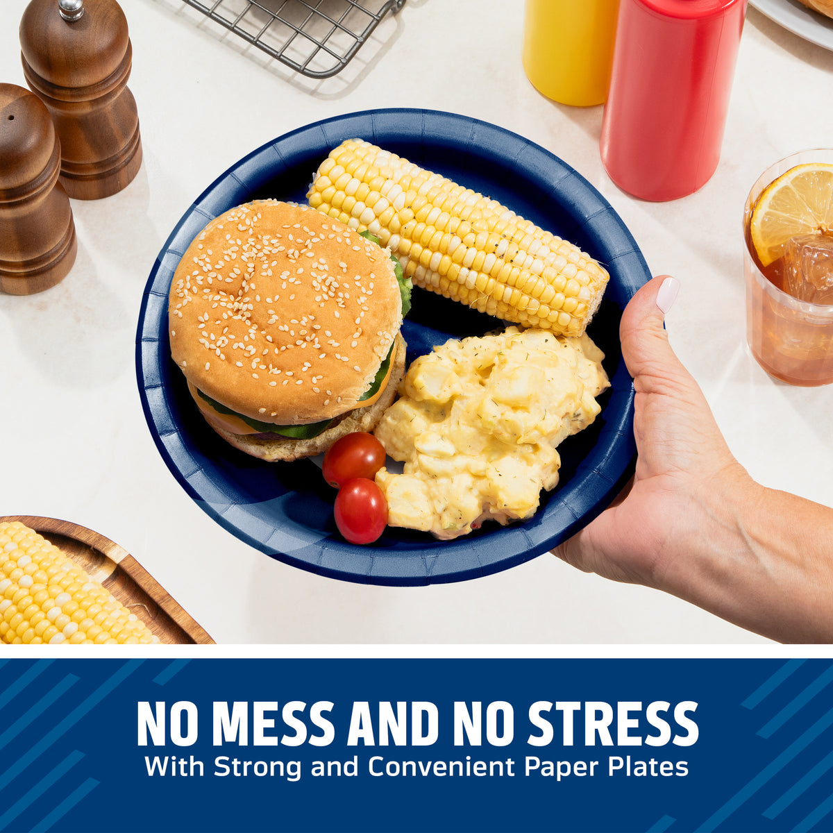 A navy blue paper plate being held by a hand, loaded with a hamburger, corn on the cob, mashed potatoes, and cherry tomatoes, set on a white countertop with condiment bottles and kitchen items in the background. A blue banner at the bottom reads 'NO MESS AND NO STRESS With Strong and Convenient Paper Plates'.