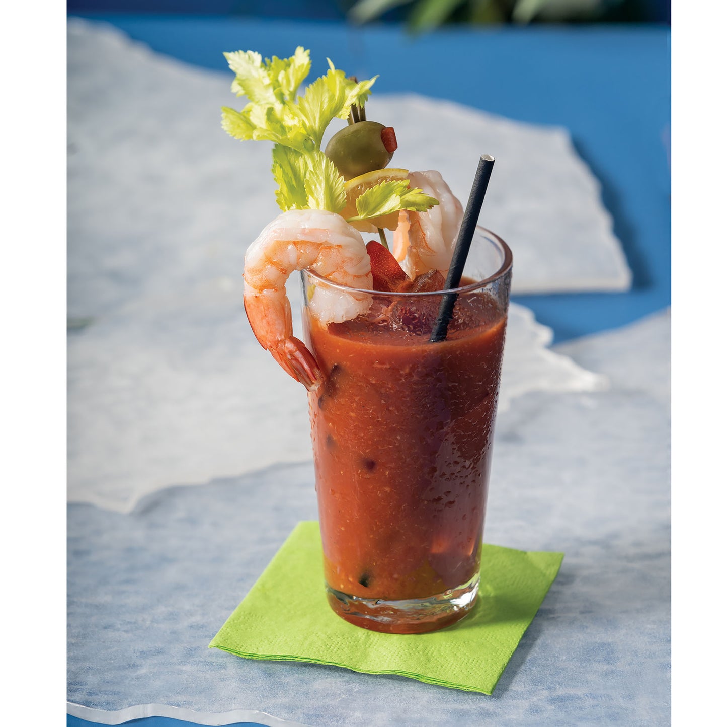 Glass of Bloody Mary with shrimp garnish and a black paper straw on a blue surface 