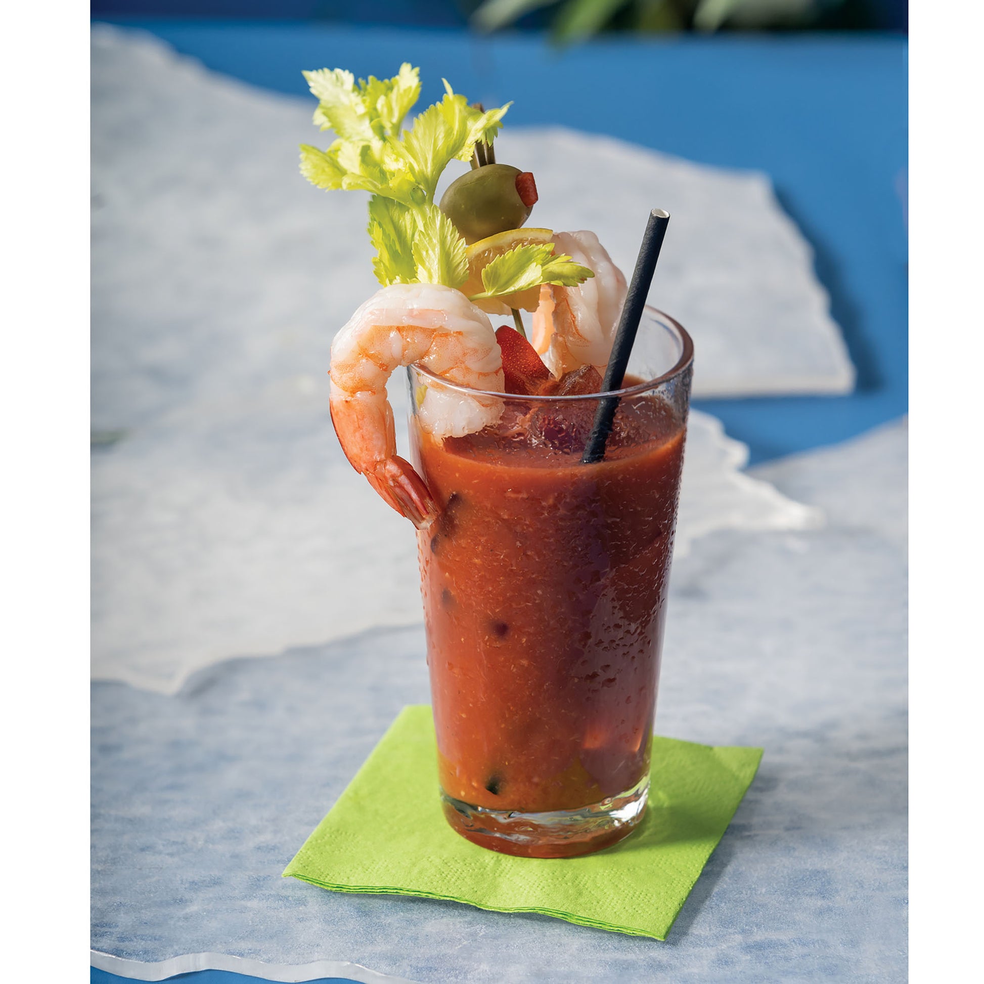 Glass of Bloody Mary with shrimp garnish and a black paper straw on a blue surface 