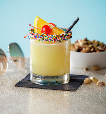 Colorful cocktail with a cherry, orange slice, and sprinkles with a black cocktail paper straws on a light blue background