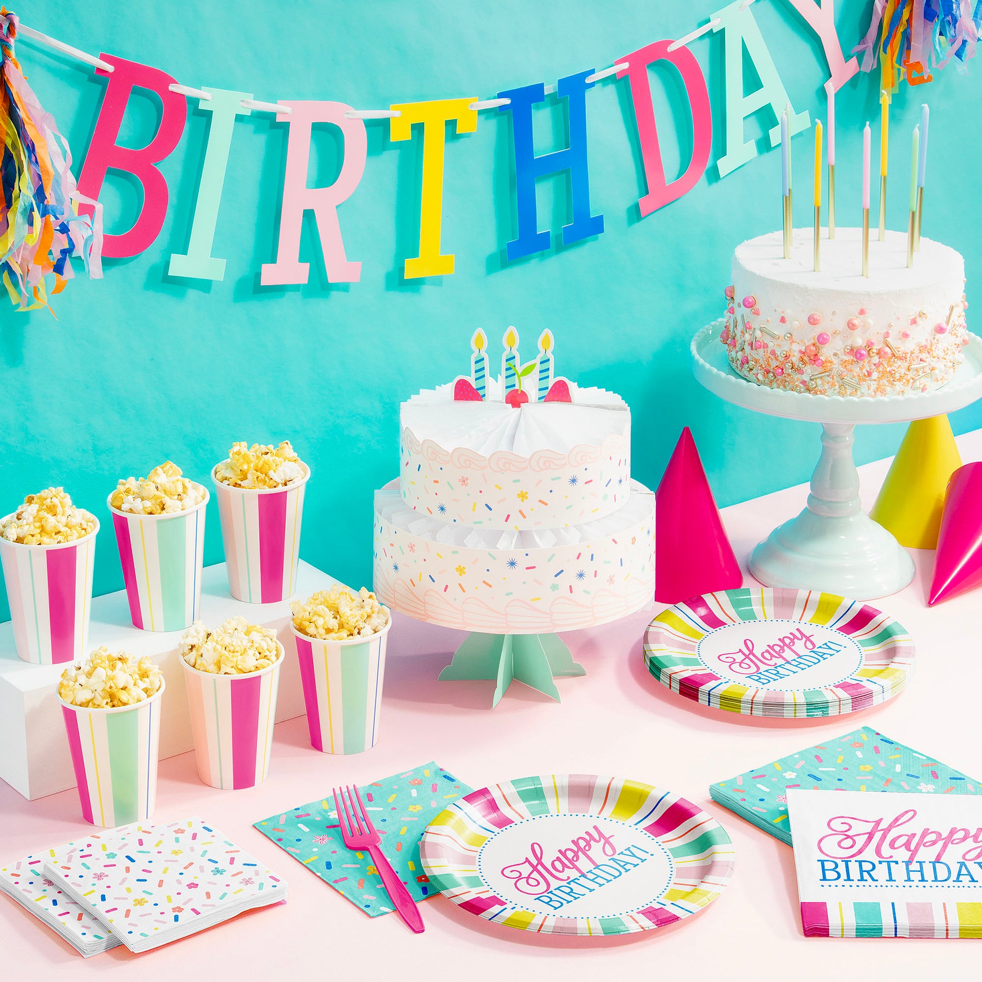 Colorful birthday tablescape with plates, cups, napkins, silverware, birthday banner and cake with candles on a light pink tablecloth