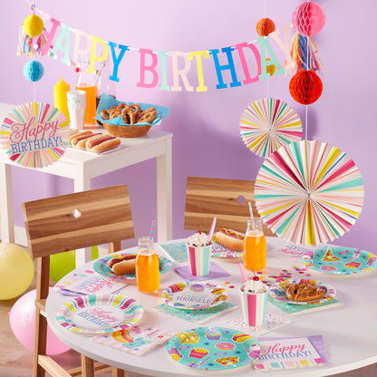 Birthday party setup with colorful decorations, tableware, and food against a purple wall.